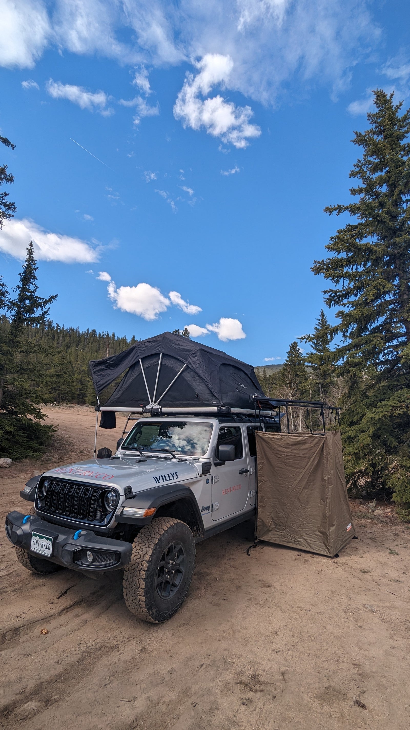 RV rental Jeep with rooftop tent & privacy shelter for off-road camping adventures under a blue sky.