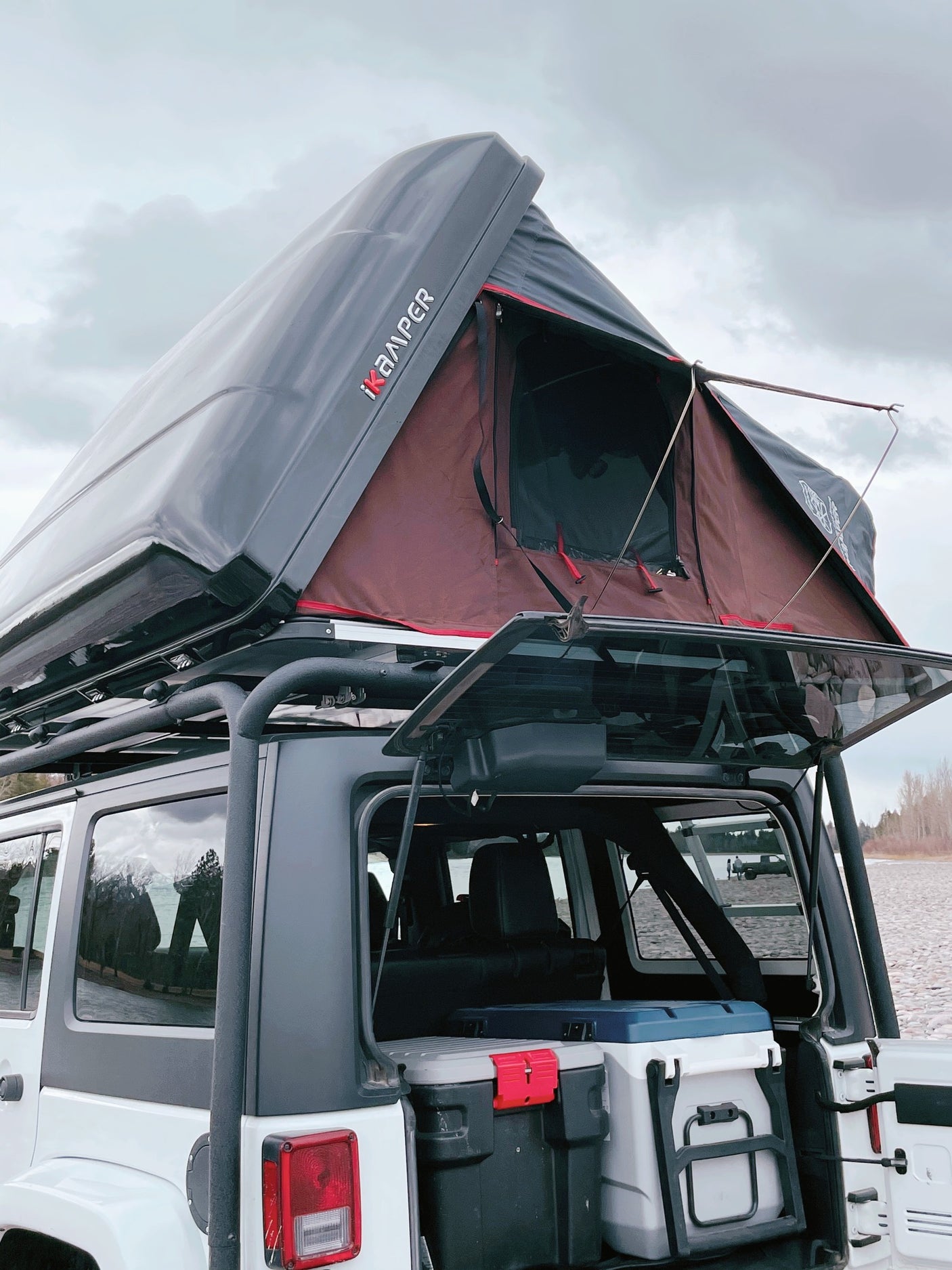 An RV rental showing a Jeep with a deployed iKAMPER rooftop tent, packed with coolers for overlanding by the lake.