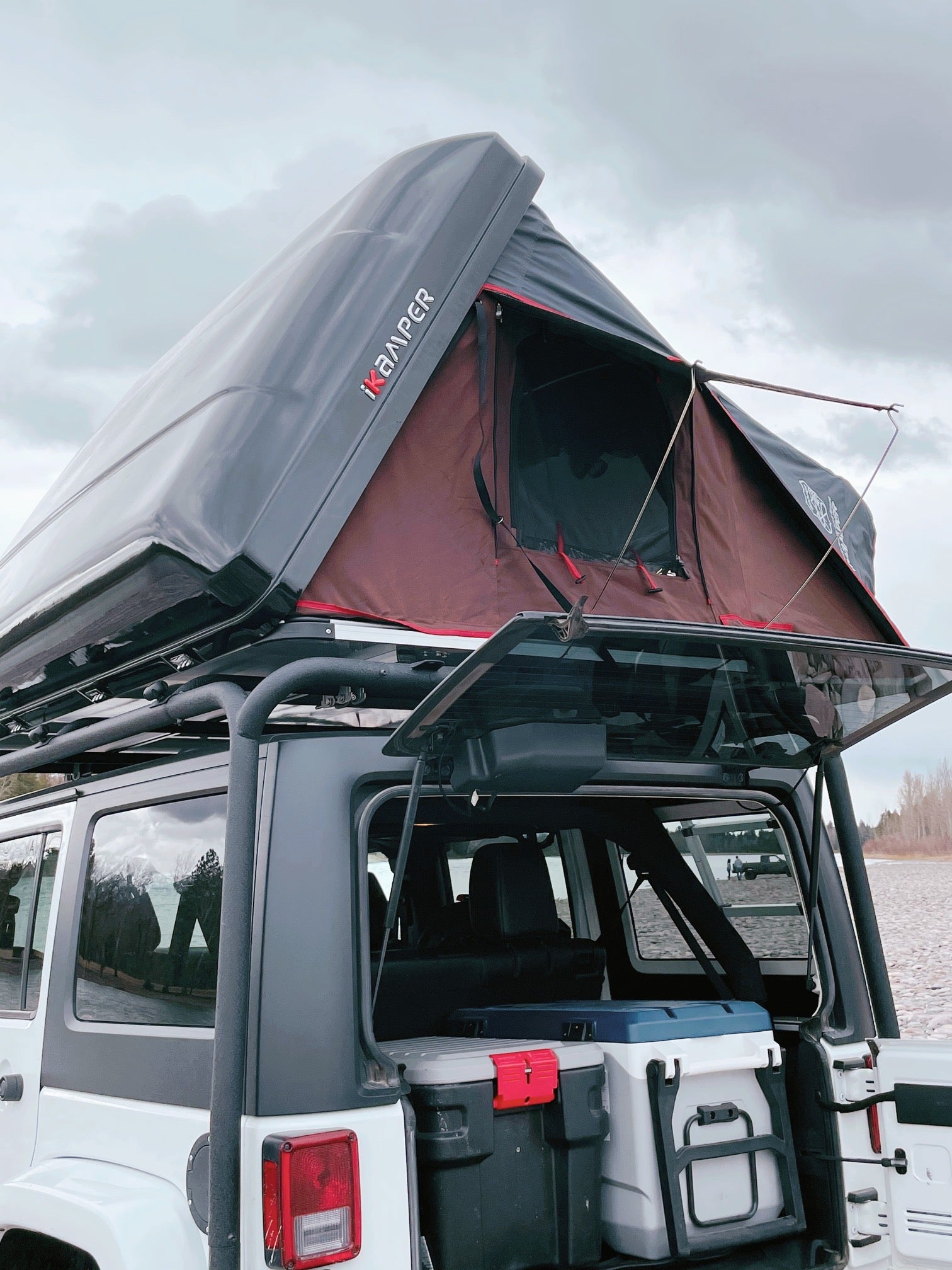 An RV rental showing a Jeep with a deployed iKAMPER rooftop tent, packed with coolers for overlanding by the lake.
