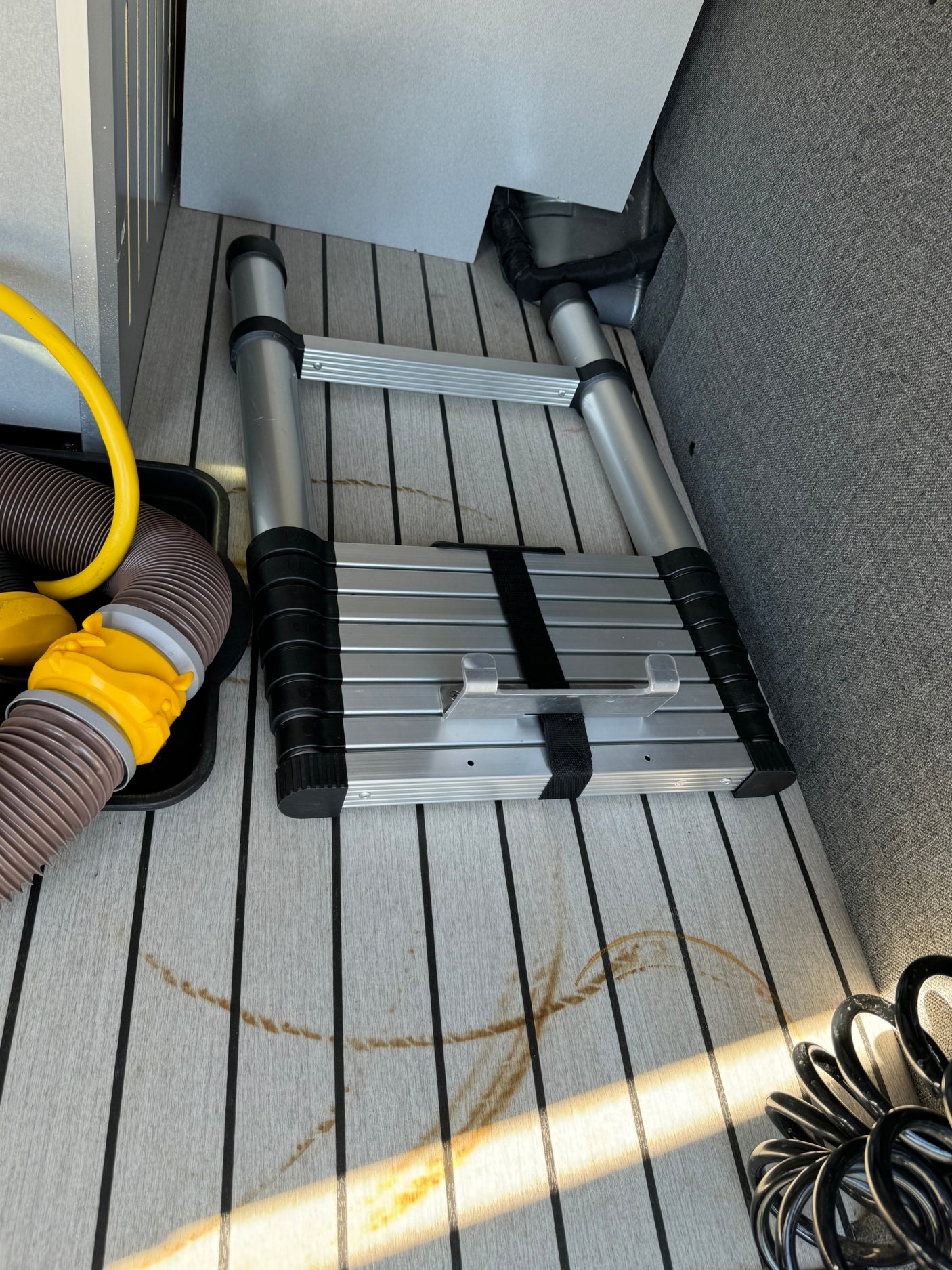 Interior RV rental storage: collapsed ladder, sewage/water hoses, and stains on faux-teak flooring.
