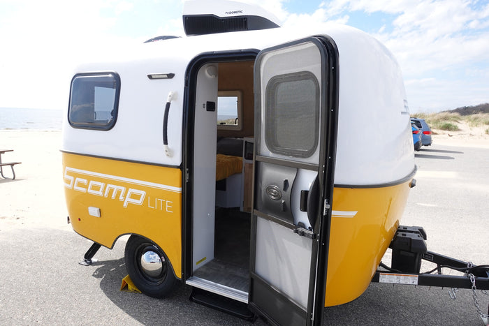 This Scamp Lite travel trailer RV rental is parked by the beach, its open door revealing a simple, cozy interior.