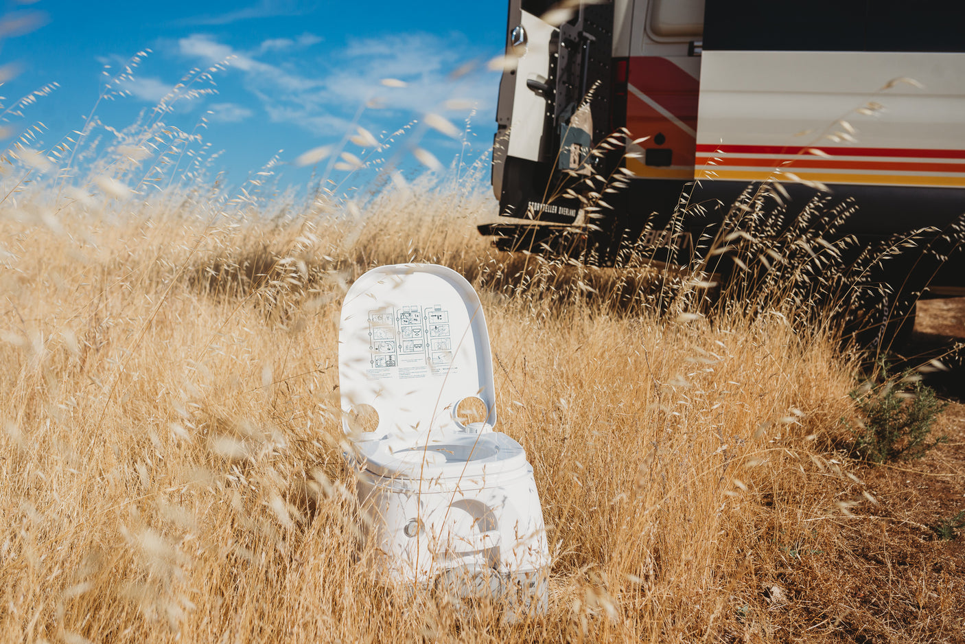 Experience RV rental freedom! Portable toilet amidst golden dry grass, with a camper van parked behind for off-grid comfort.