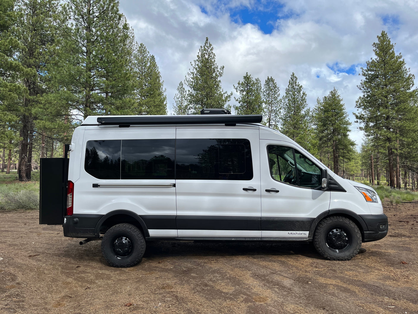 White Ford Transit rv rental camper van parked in a forest with pine trees under a cloudy sky, ready for adventure.