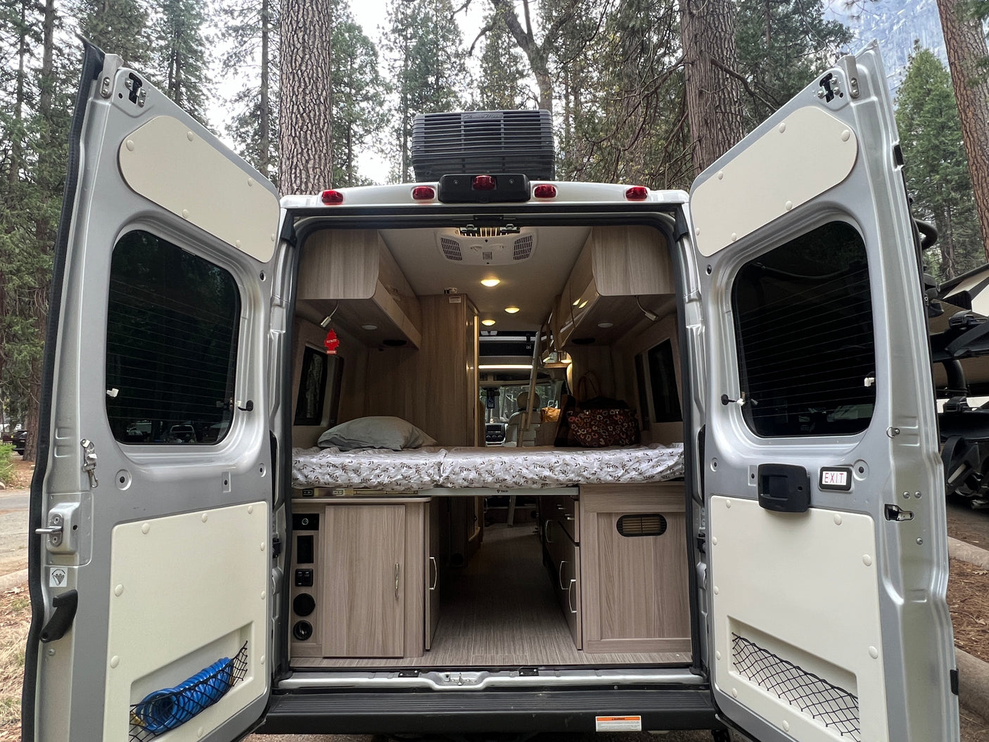 Rear view of an RV rental camper van, doors open to reveal a comfortable bed, light wood cabinetry, and integrated controls.