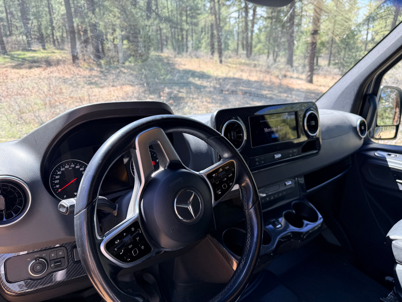 Interior of a Mercedes-Benz RV rental, showing the steering wheel, dashboard, and a forest view outside the windshield.