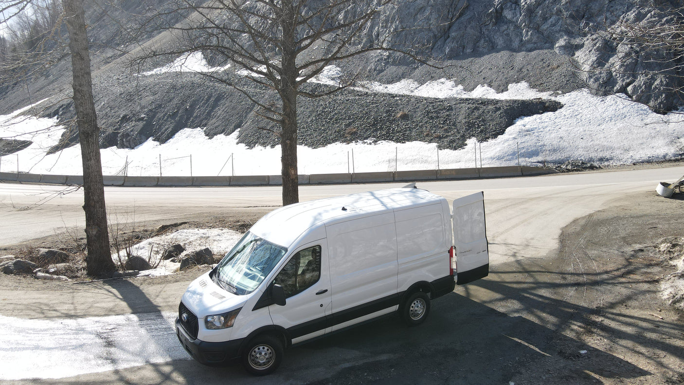 White van, like an RV rental, parked by a snowy mountain road with bare trees. Rear doors are open.