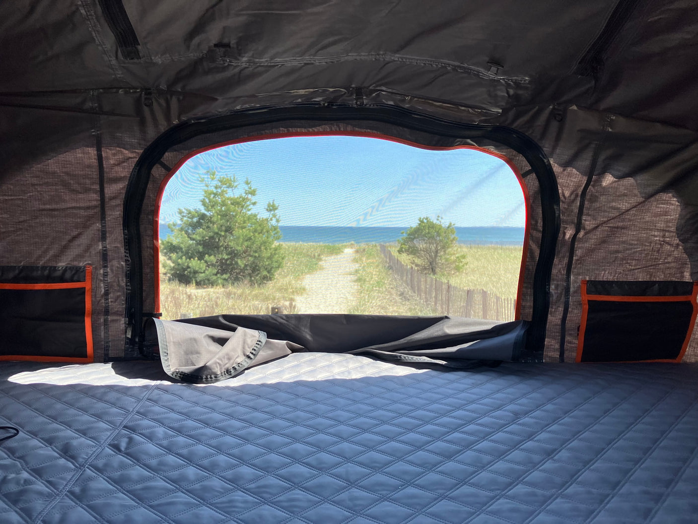 View from an RV rental: a cozy, quilted bed and interior storage, with a scenic window overlooking a sandy beach path to the ocean.
