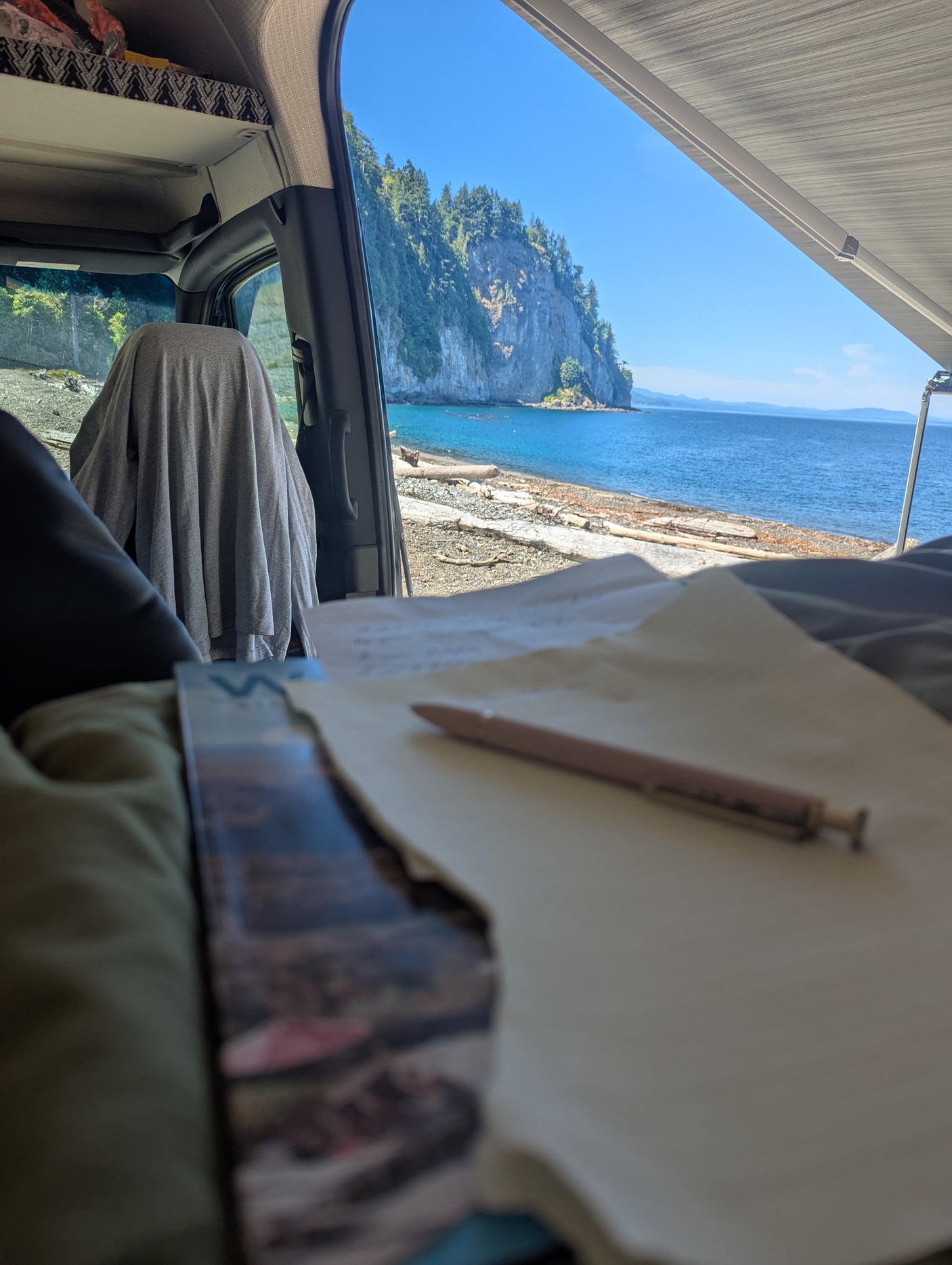 Working inside an **rv rental** camper van, overlooking a stunning cliff-lined beach and serene blue ocean under an extended awning.