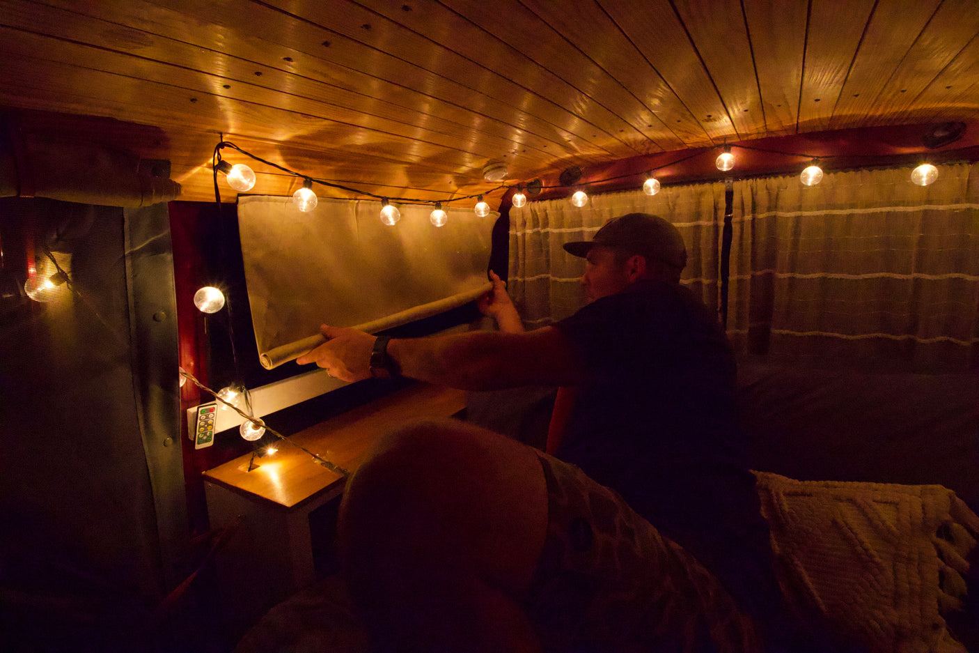 Enjoy a cozy rv rental experience! Man adjusts window shade inside a warmly lit camper van with string lights.