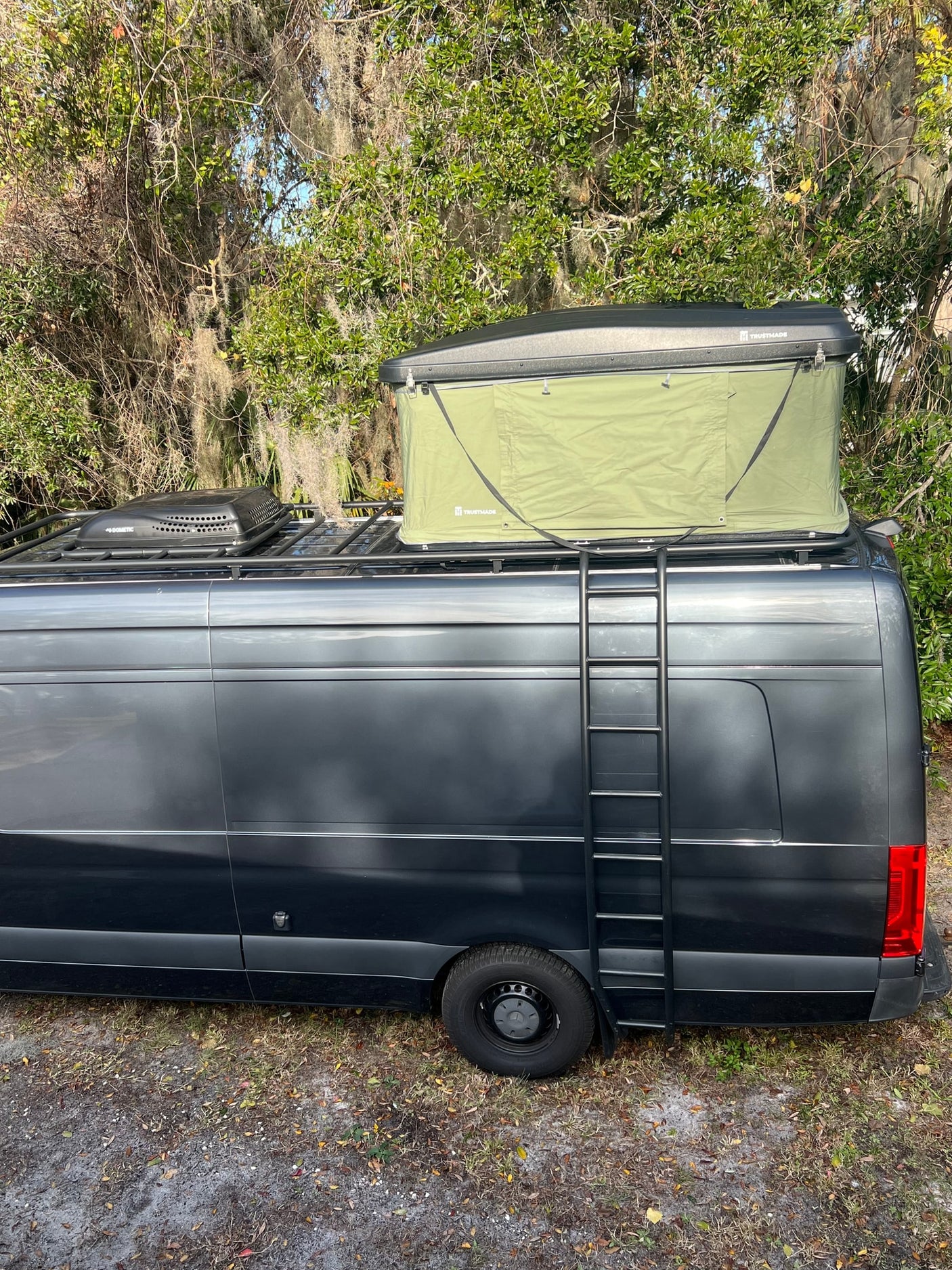 This dark gray RV rental camper van, equipped with a Trusthmade rooftop tent and Dometic unit, has a side ladder.