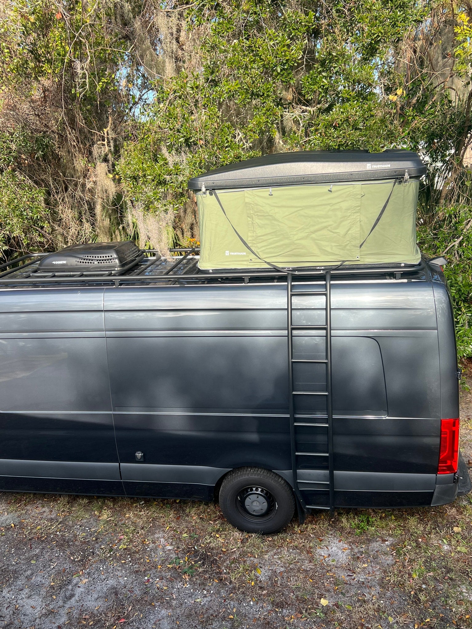 This dark gray RV rental camper van, equipped with a Trusthmade rooftop tent and Dometic unit, has a side ladder.