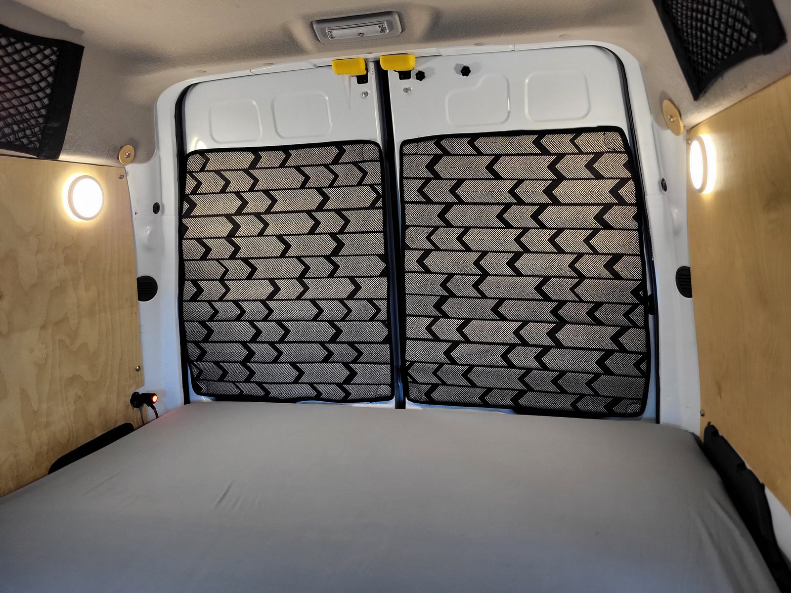 Spacious RV rental camper van interior: bed, custom patterned rear door window covers, and practical lighting on wood panels.