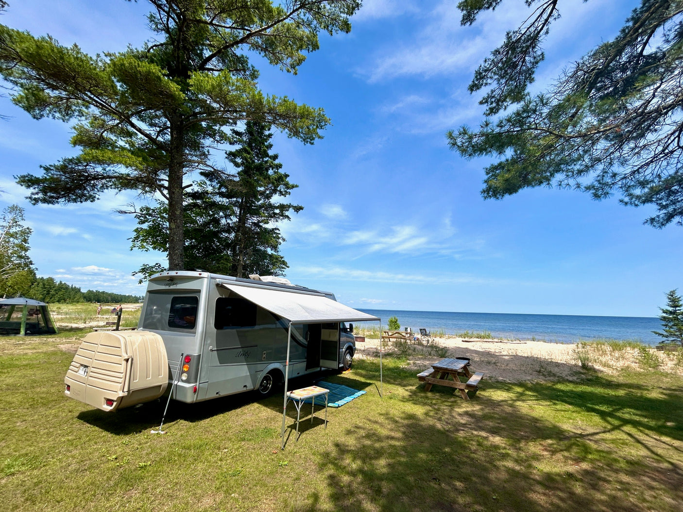 Scenic rv rental parked beachfront. Gray RV with awning, picnic table, and storage next to sand, water, and trees under blue sky.