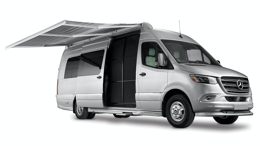 and a forest leading to blue water.|Enjoy nature with an RV rental! A couple walks in a grassy field beside their silver camper van with an open door.|This silver Mercedes-Benz Sprinter rv rental van shown with an extended awning and open side door