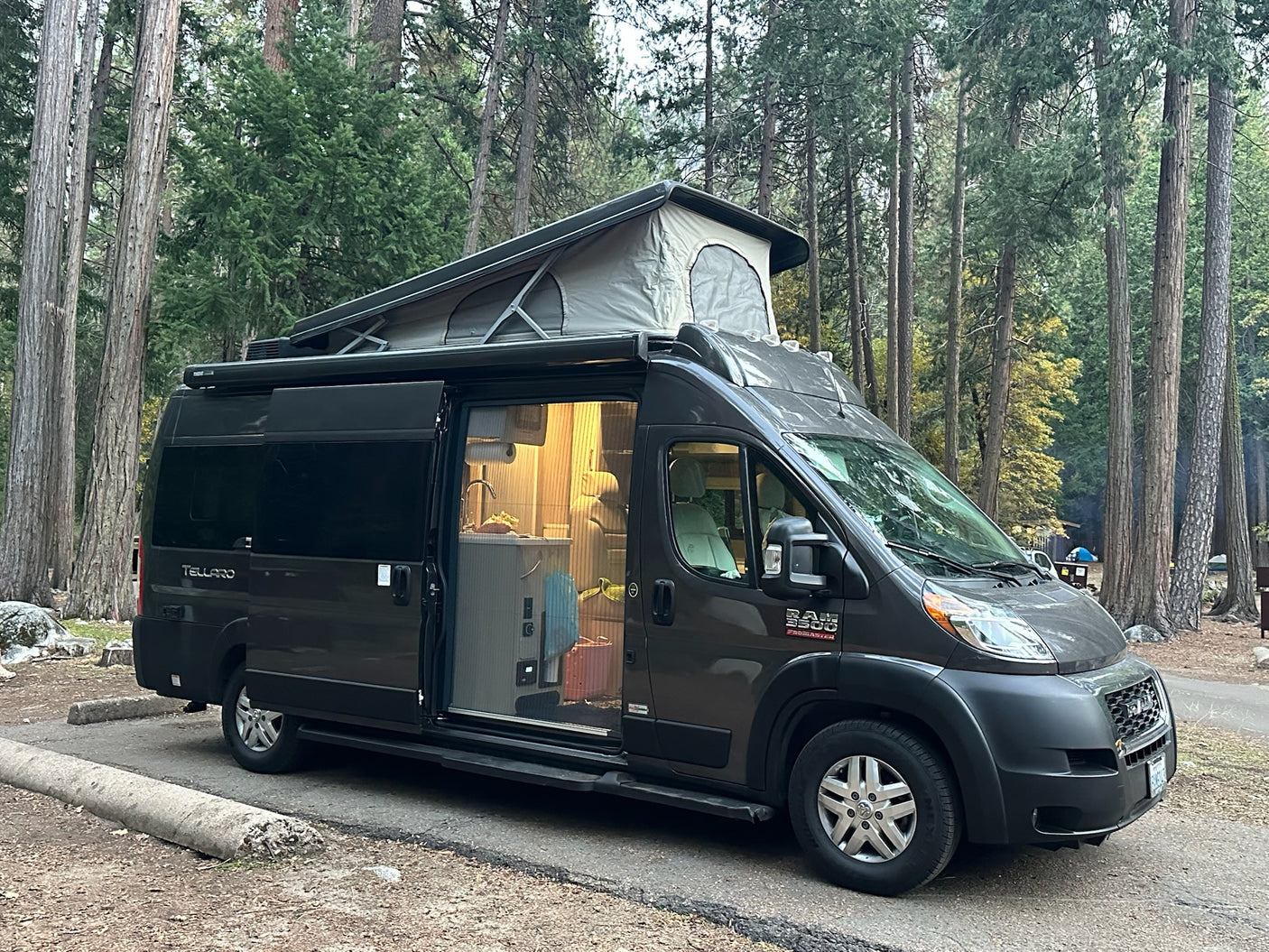 Experience nature in this gray pop-top rv rental, parked among towering trees, with its interior lit for a cozy campsite stay.