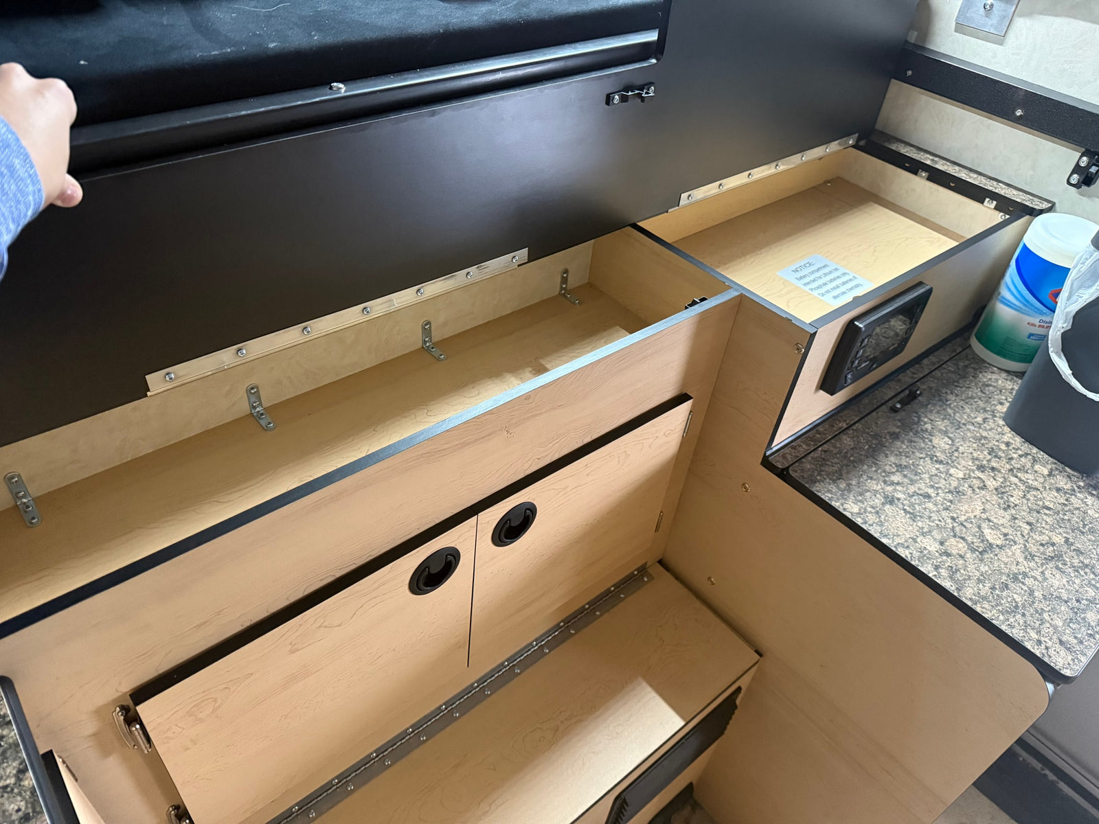 Inside an RV rental: open wooden storage compartments, black panels, and a counter with a battery notice, showing efficient interior design.