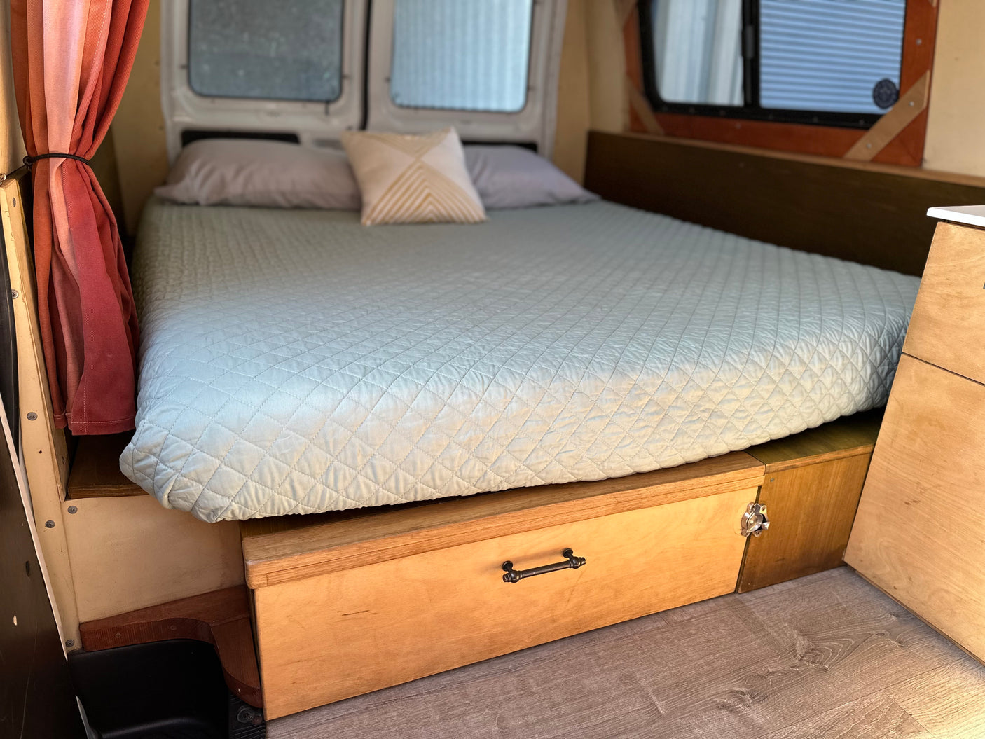 RV rental interior: A cozy bed with a light blue quilted cover and a decorative pillow. Underneath are wooden storage drawers.