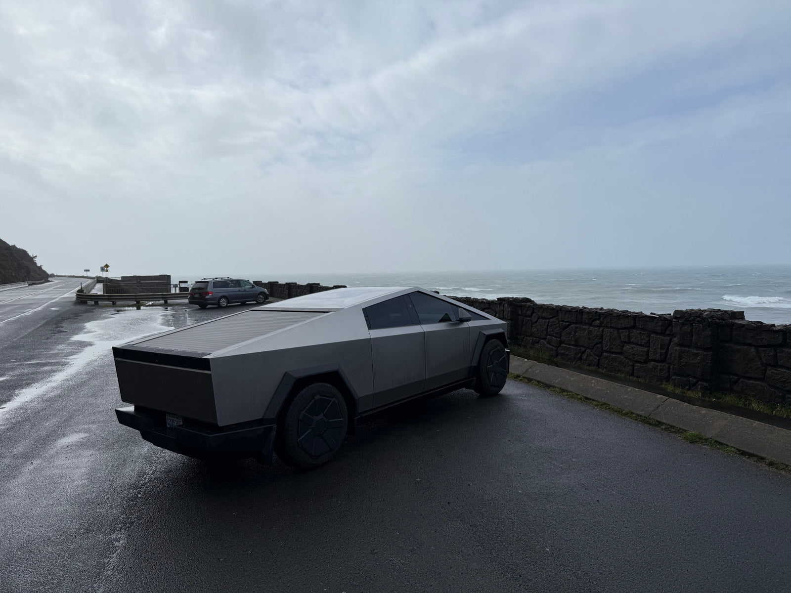 Sleek Tesla Cybertruck on a wet coastal road overlooking a stormy ocean. Forget an RV rental, this is modern exploration.