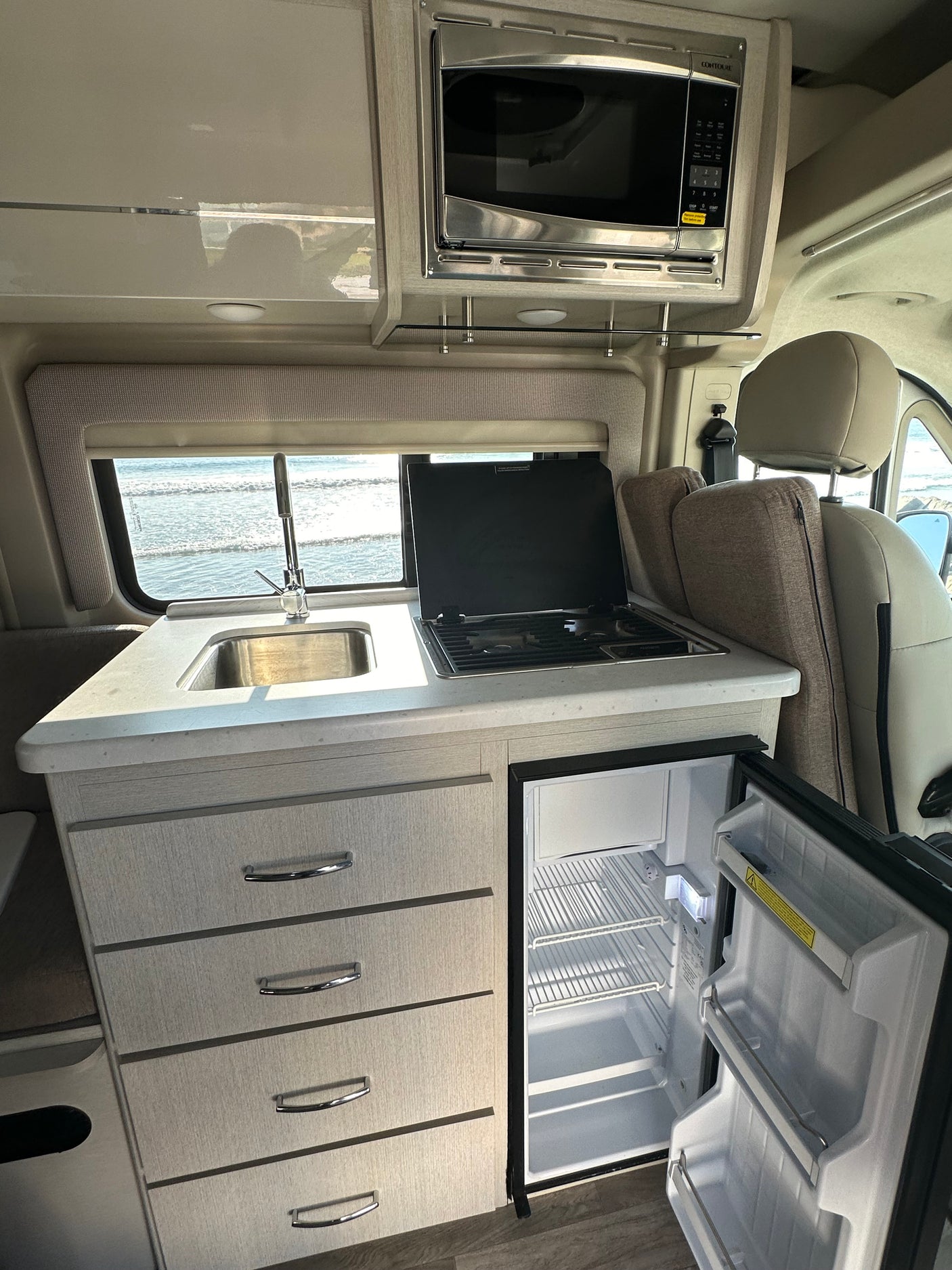 Inside this RV rental, a compact kitchen with sink, stovetop, microwave, and open fridge faces an ocean view.