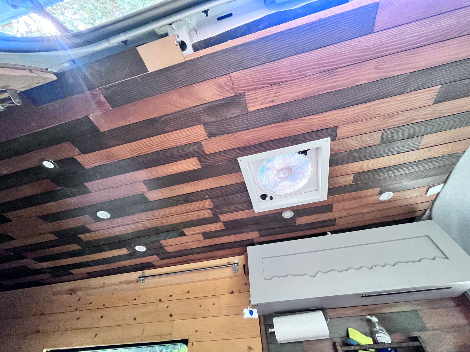 See a cozy RV rental interior: alternating wood ceiling planks, bright lights, central fan, and wall-mounted storage.