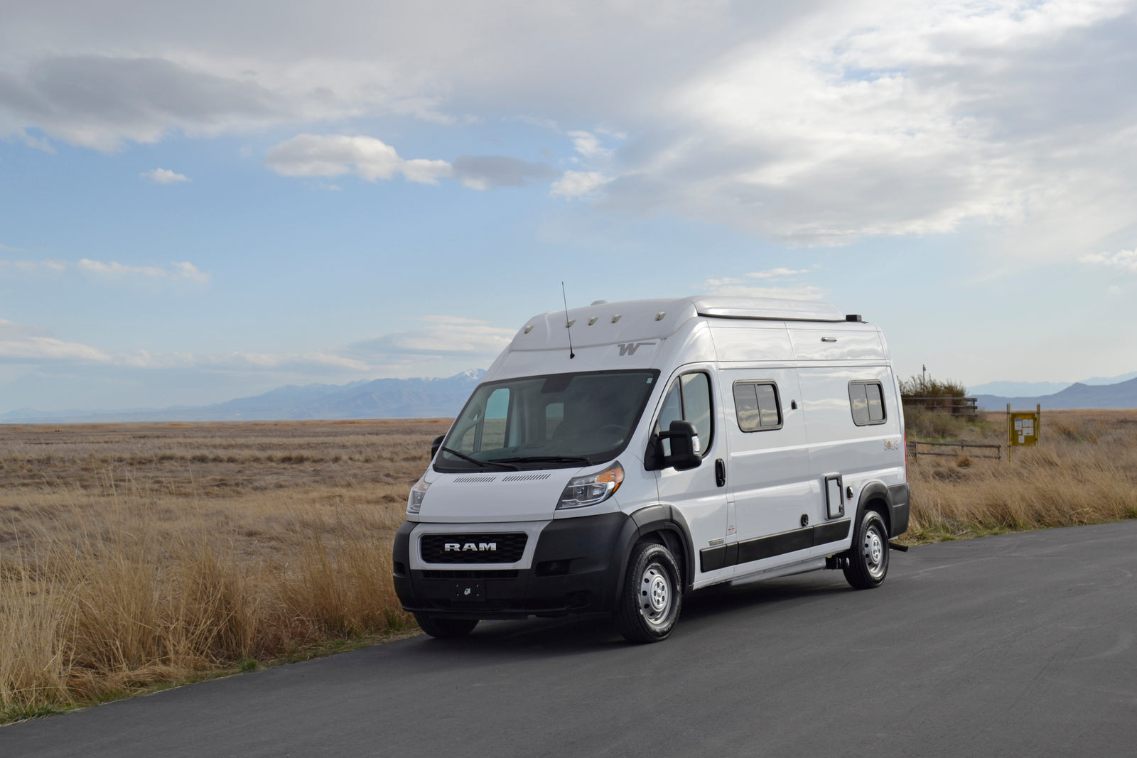 This white RV rental van is parked on an asphalt road, set against a vast landscape of dry fields and distant mountains under a cloudy sky.