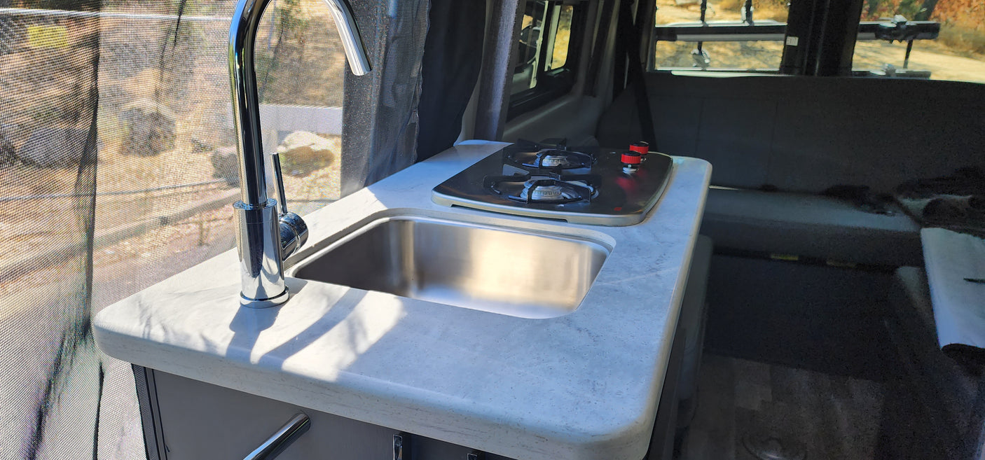 Compact kitchen in an RV rental, featuring a chrome faucet, stainless steel sink, and two-burner stove by a screened window.