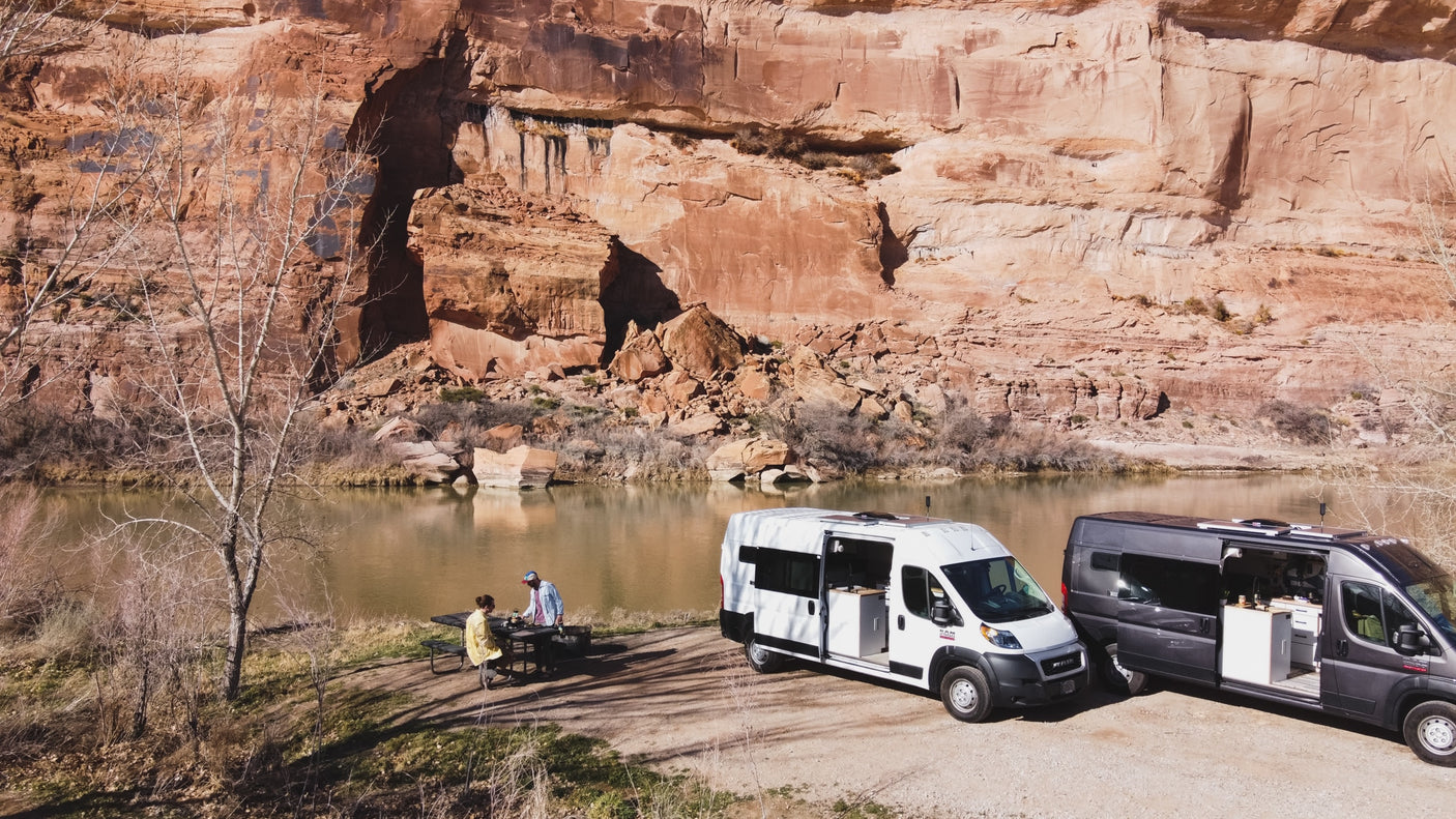 Scenic riverfront camping: Two camper van RV rentals (white & grey) by a river with red cliffs. People picnic outdoors.
