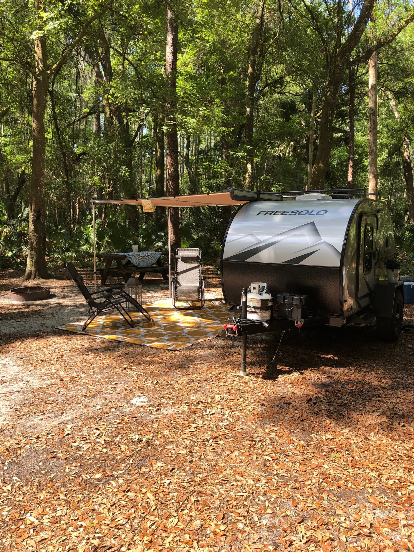 An RV rental camper with an awning, picnic table, and chairs set up in a serene, leafy forest campsite on a sunny day.