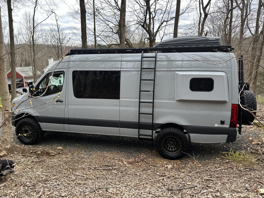 Gray Sprinter camper van, ideal for rv rental adventures, featuring a roof rack, ladder, and off-road tires, parked outdoors.