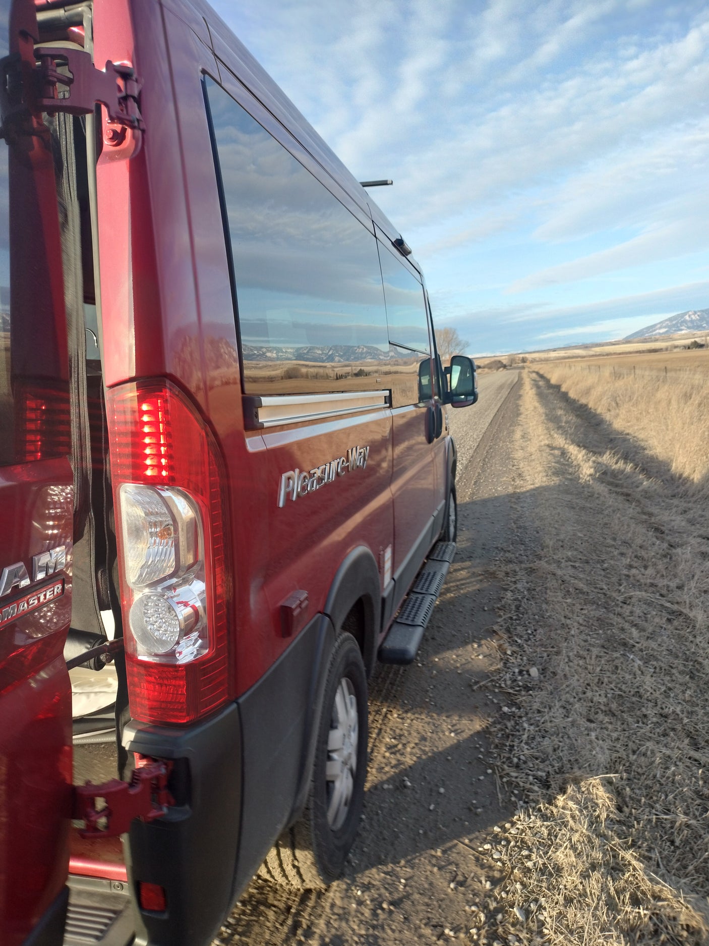 Scenic rv rental: Red Pleasure-Way camper van on a dirt road. Rear doors open, mountains reflected in side windows.