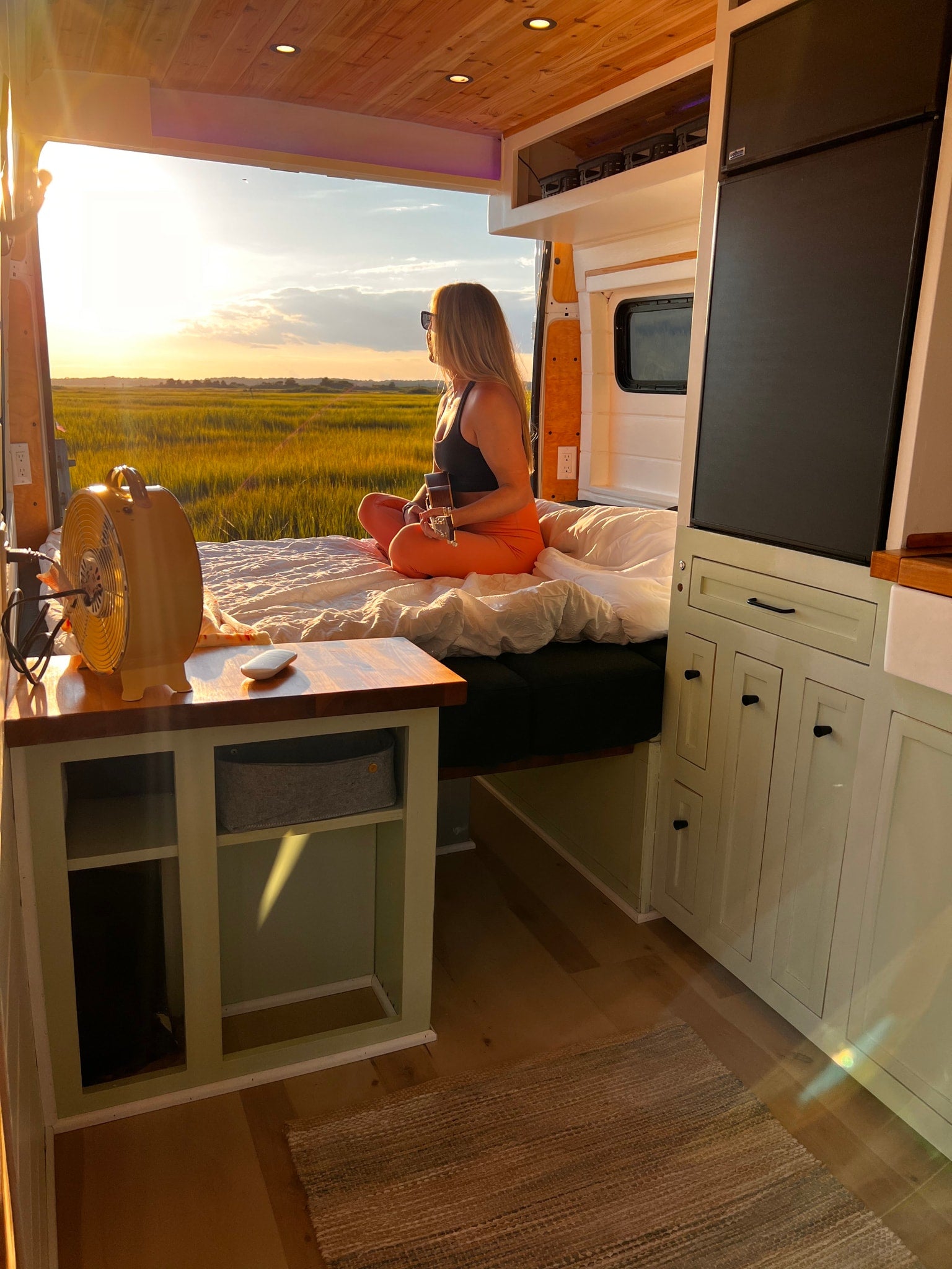 Relaxing RV rental: A woman plays ukulele in a cozy camper van, enjoying a golden sunset over a marsh.