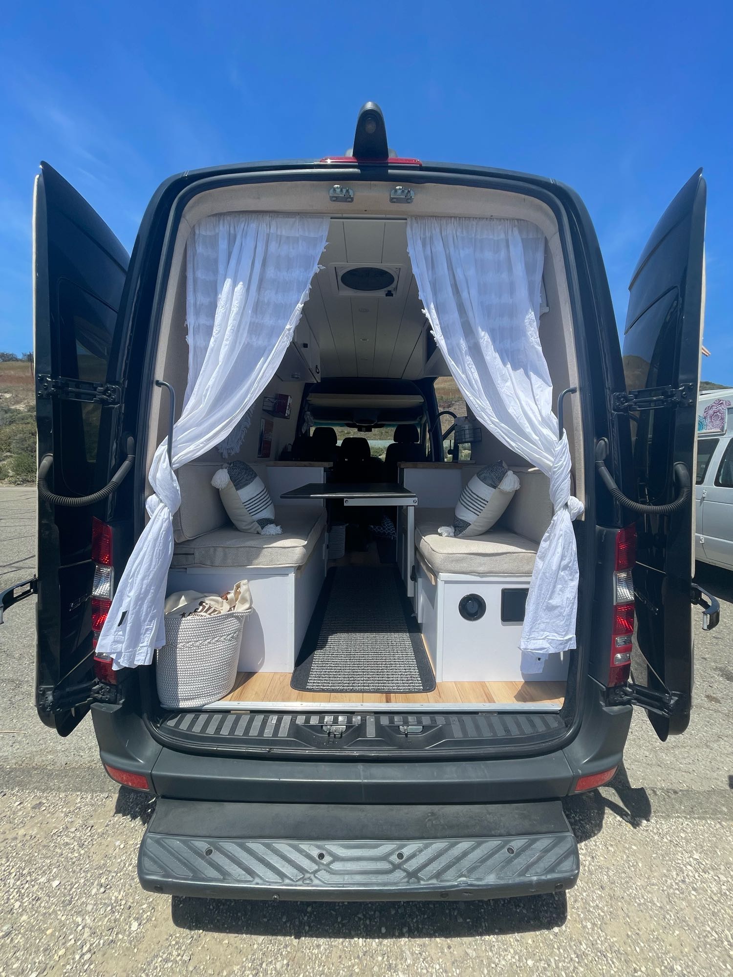 Open back of a black RV rental, revealing a cozy camper van interior with bench seats, table, and white curtains.
