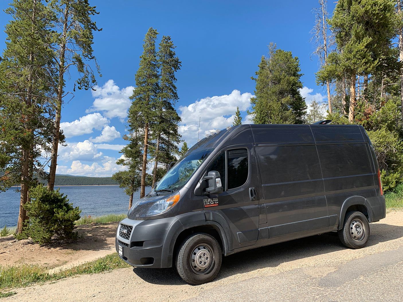 Dark gray rv rental camper van parked by a serene lake amid pine trees under a blue sky with white clouds.