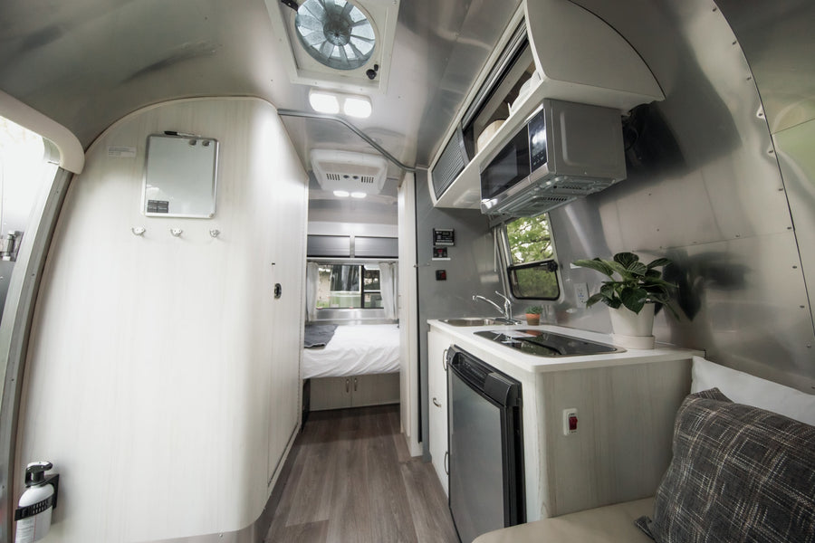 Modern RV rental interior: kitchen with microwave, fridge, sink; cozy bedroom; sleek metallic and light wood paneling throughout.