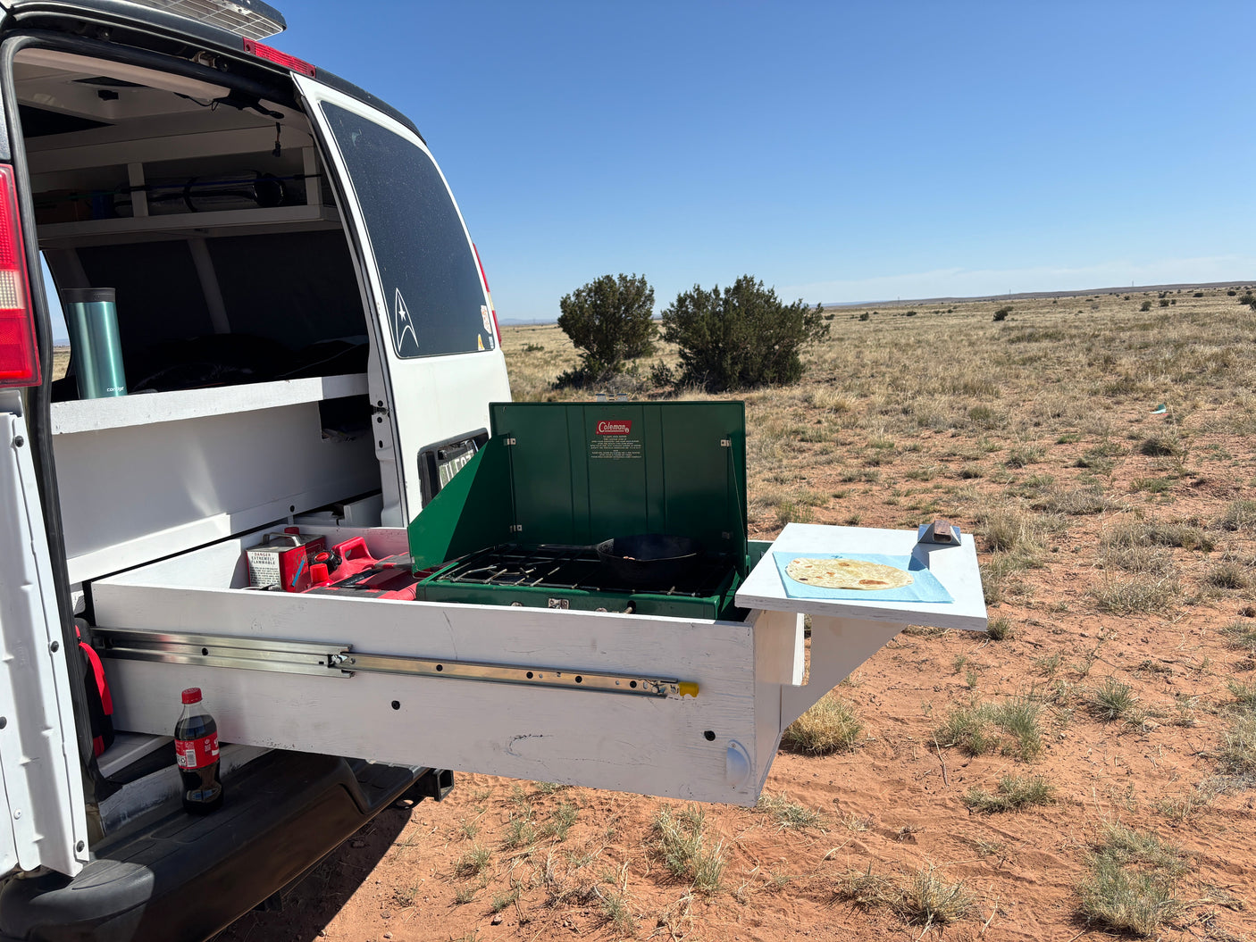 RV rental outdoor kitchen: A pull-out stove from a camper van cooking in an arid landscape with tortillas on a side table.