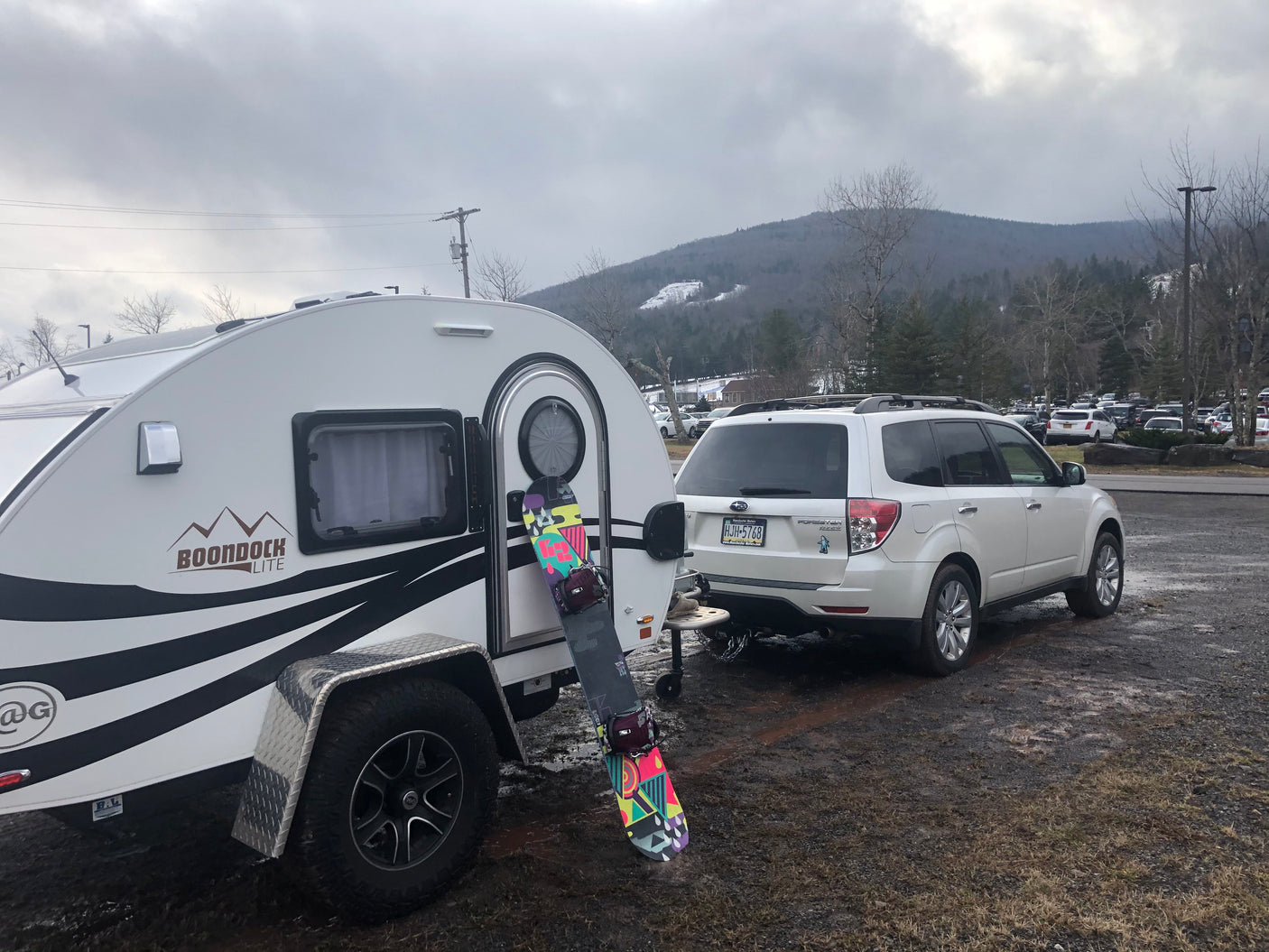 White Boondock Lite teardrop camper (RV rental) and Subaru Forester, with a snowboard, at a snowy mountain resort.