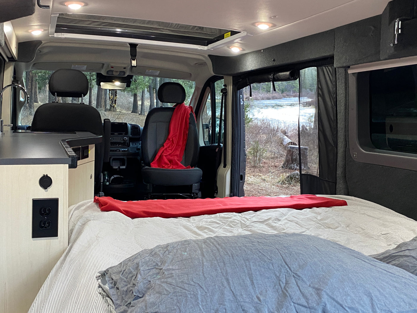 Cozy RV rental interior with a comfortable bed, kitchenette, and a peaceful view of a wintry river and trees outside.