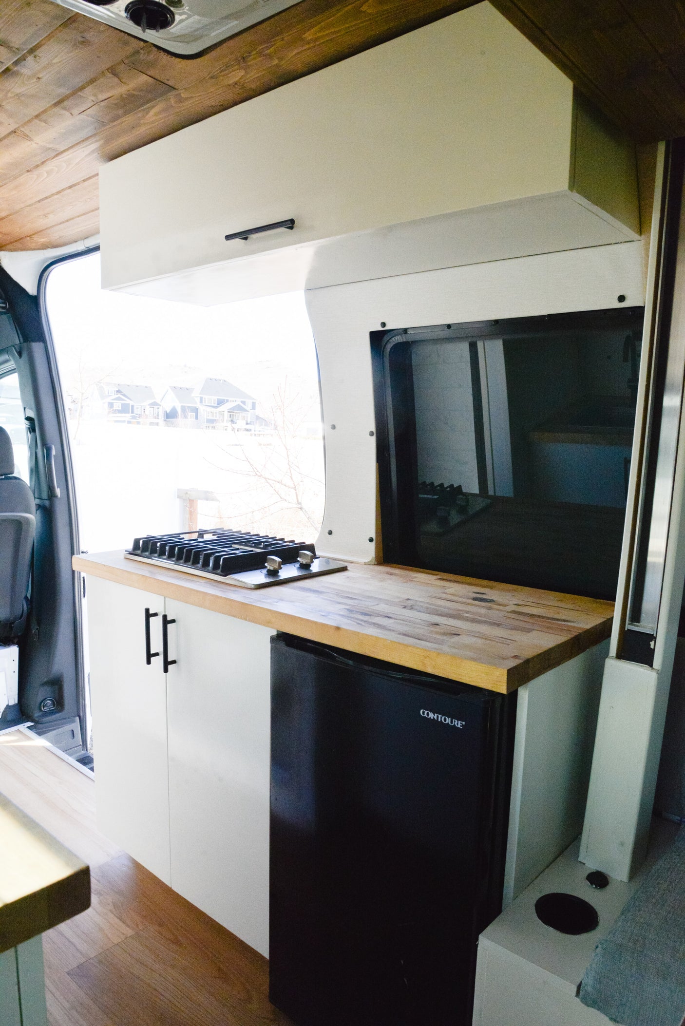 RV rental kitchen: stylish van conversion features a gas stove, wooden counter, mini-fridge, and white overhead cabinets.