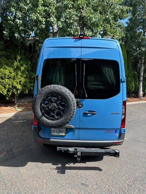 Rear view of a bright blue Mercedes Sprinter camper van, ideal for RV rental. Features a mounted spare tire and hitch.