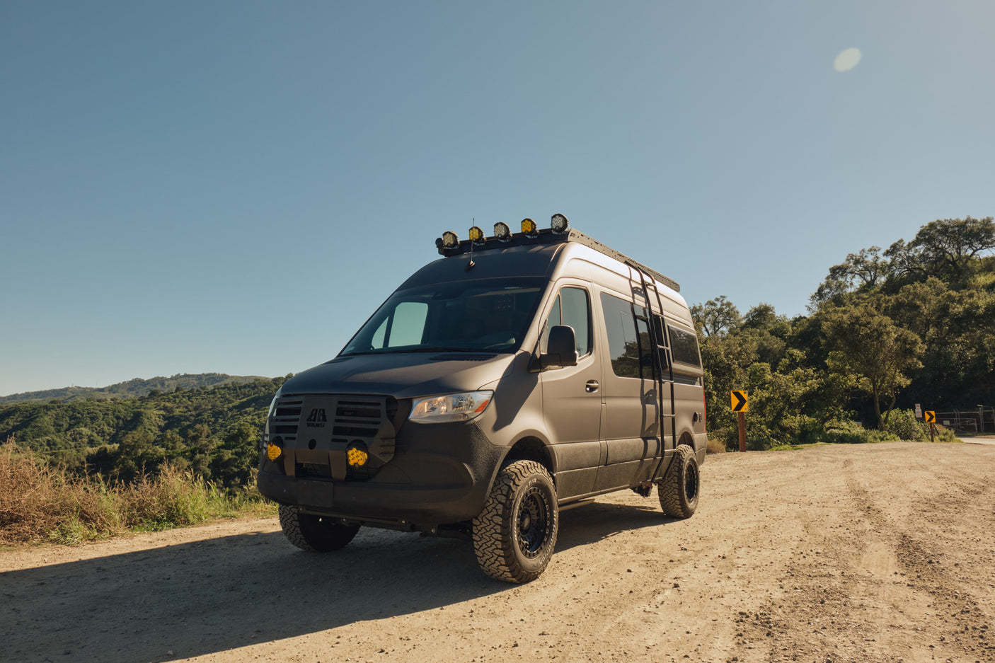 Rugged off-road Sprinter adventure van, a premium RV rental, parked on a scenic dirt road with green hills.