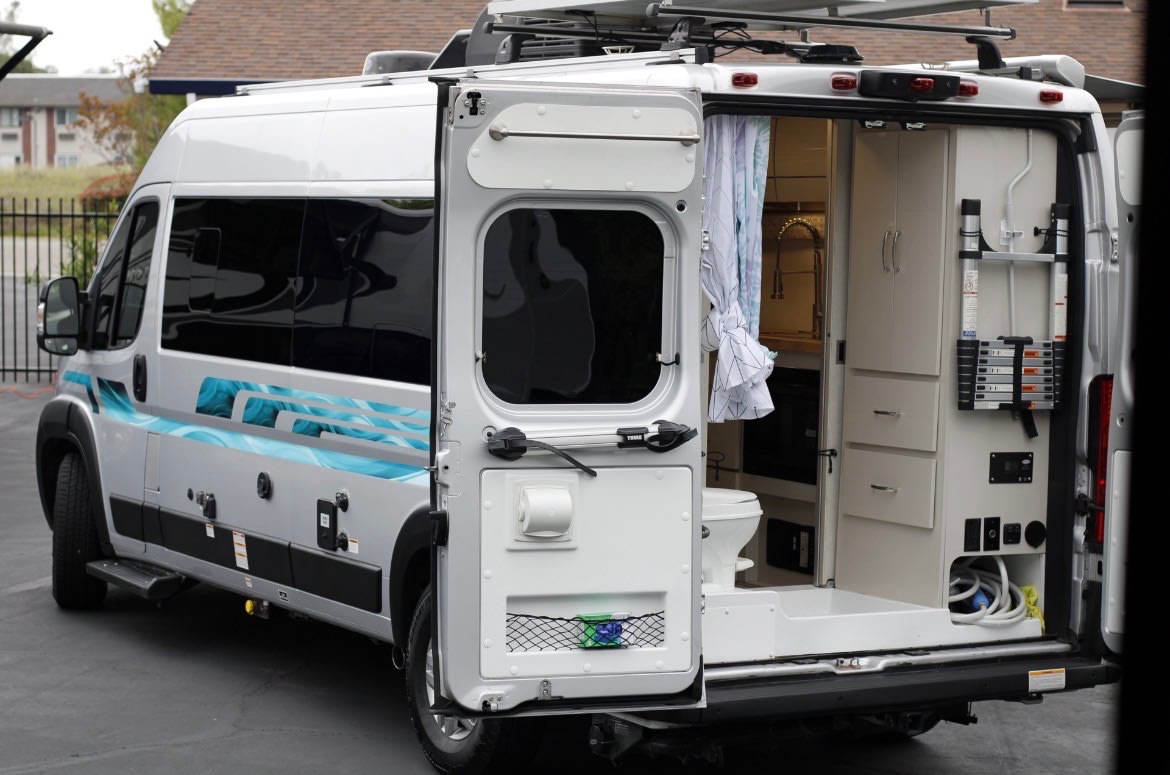 Silver RV rental van with rear doors open, showing the built-in kitchen, toilet, and storage inside, along with exterior utility hookups. — photo 3