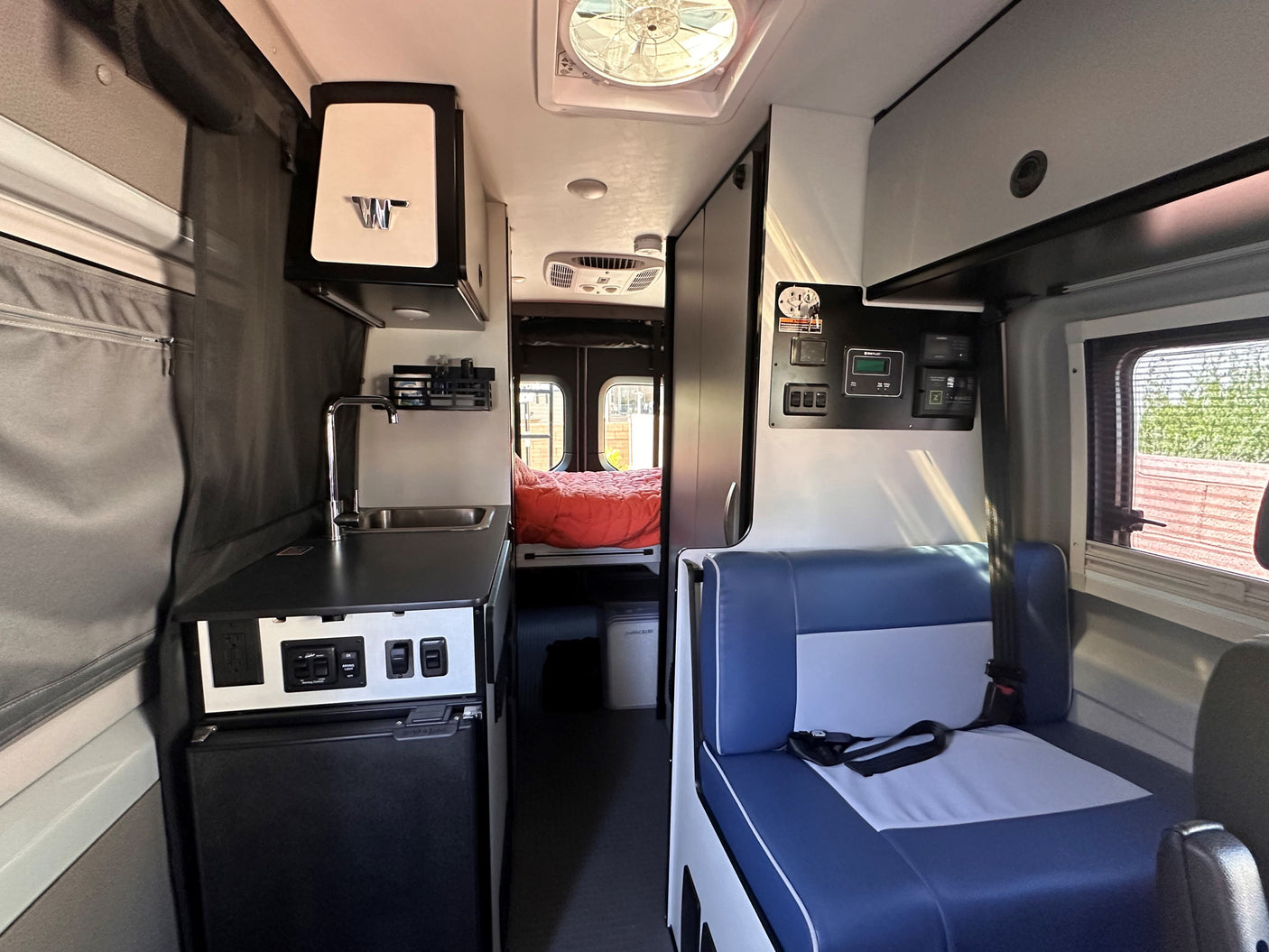Step inside this RV rental: a compact interior features a kitchenette, controls, blue seating, and a cozy bed in the rear.