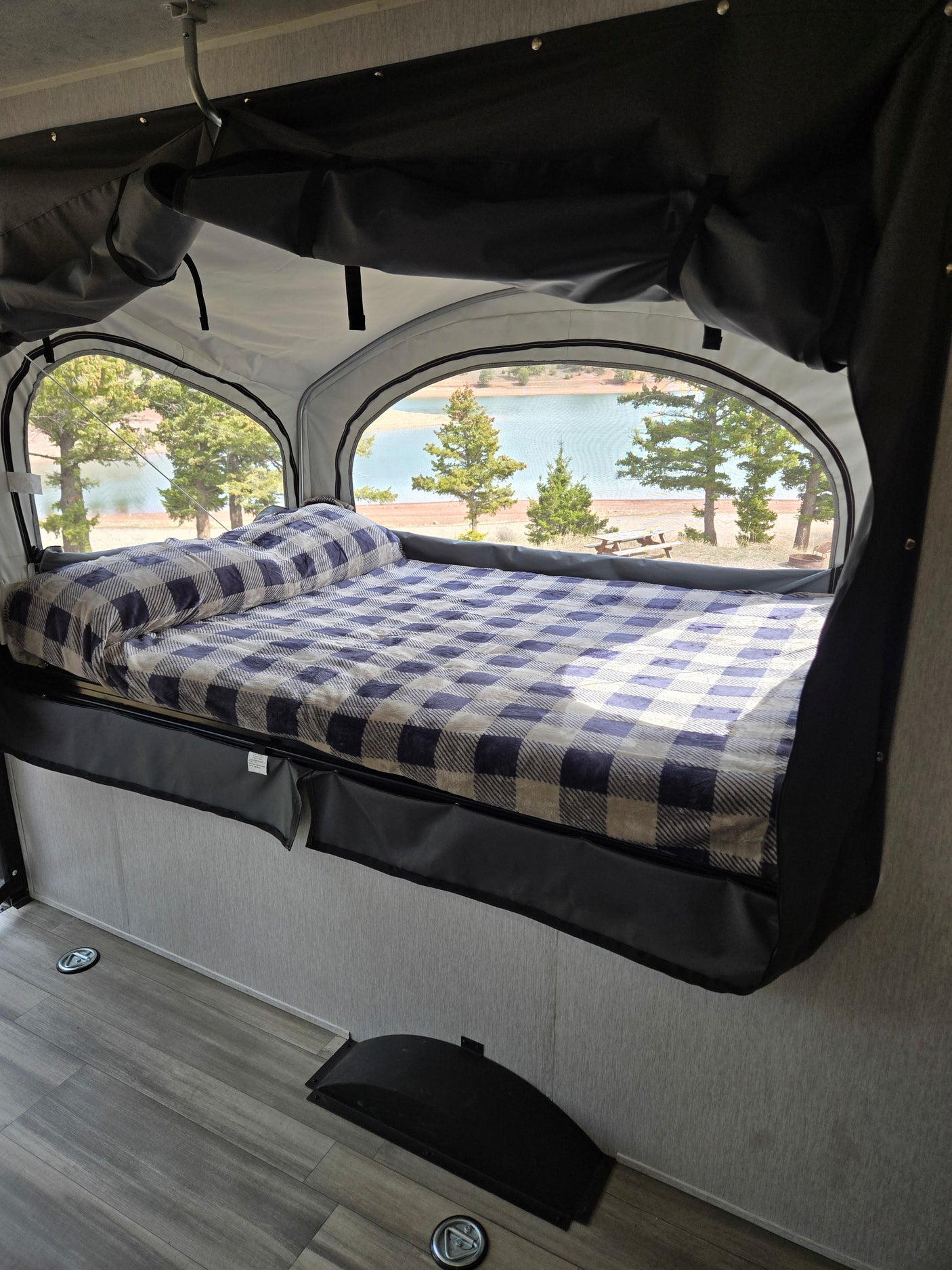 Enjoy scenic lake views from this cozy RV rental's bed with a plaid blanket, framed by arched windows and pine trees.
