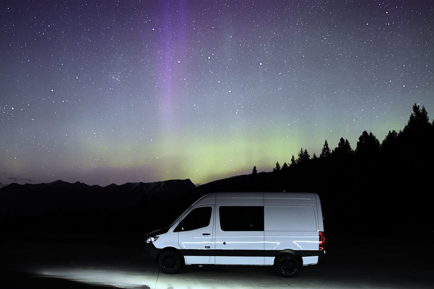 A white RV rental van illuminated under vibrant purple/green aurora, starry sky, silhouetted mountains & trees. Epic adventure awaits.