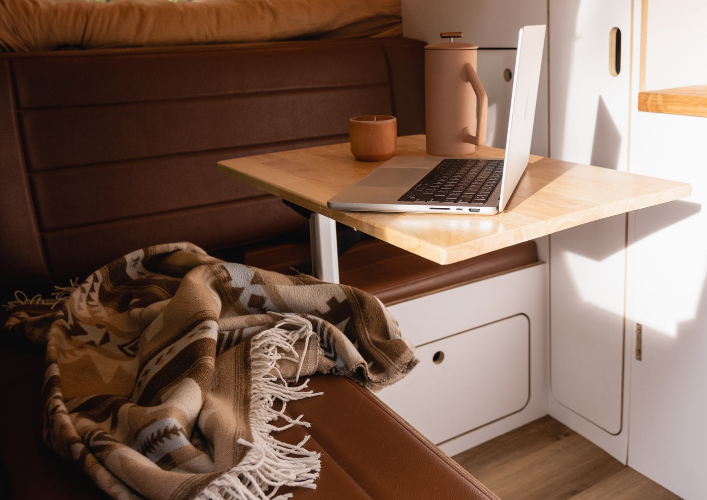 Cozy RV rental interior: laptop workstation with coffee on a wooden table, warm blanket on a brown seat.