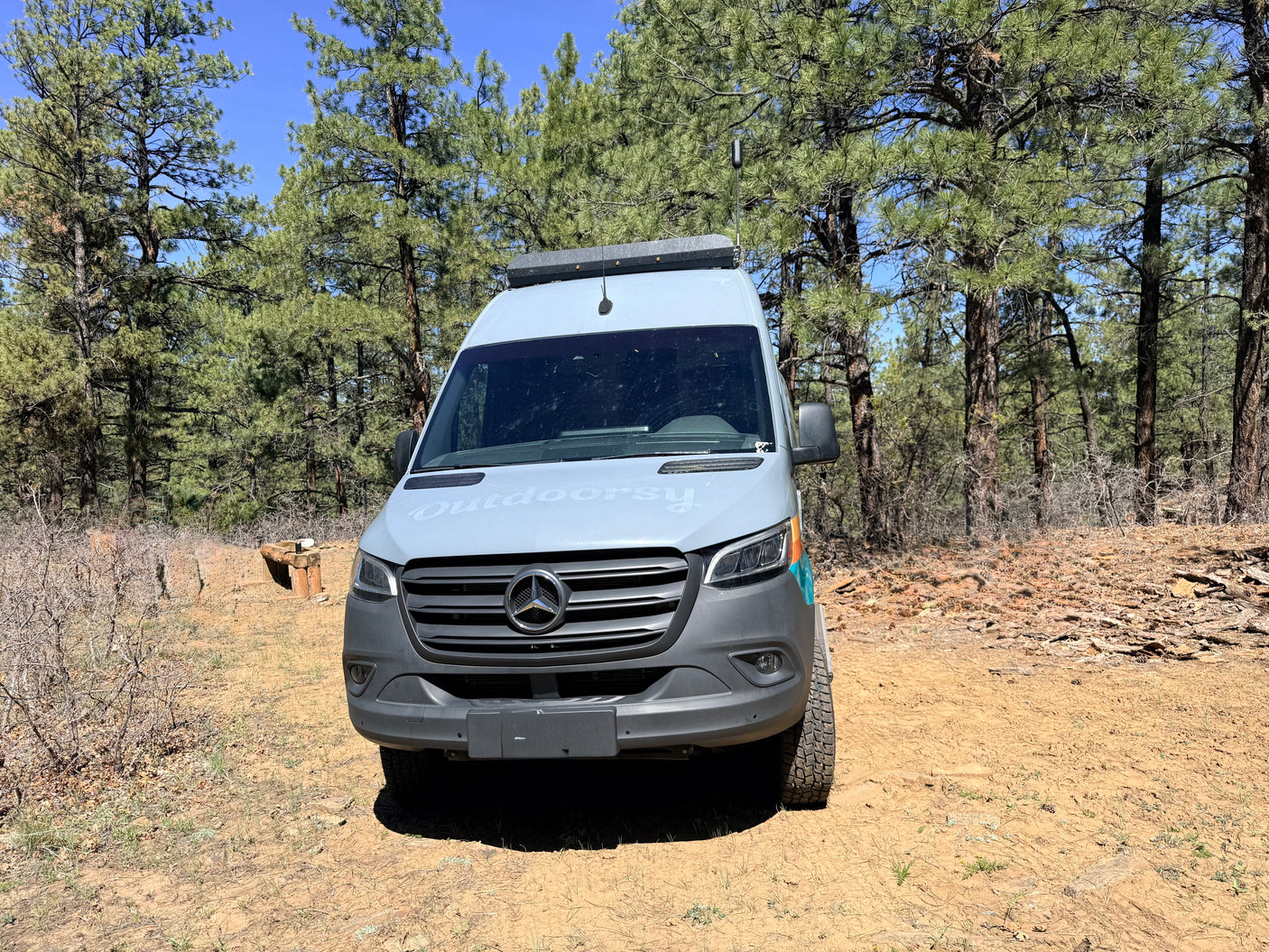 Outdoorsy RV rental: a grey Mercedes-Benz Sprinter camper van parked on a dirt road under tall pine trees.