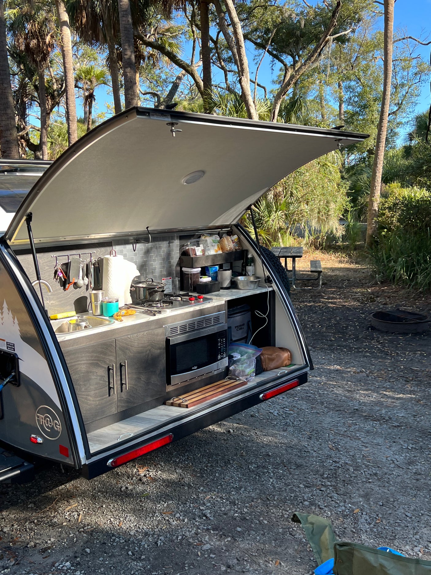 An rv rental teardrop trailer with a fully equipped outdoor kitchen, set in a sunny, treed campsite.