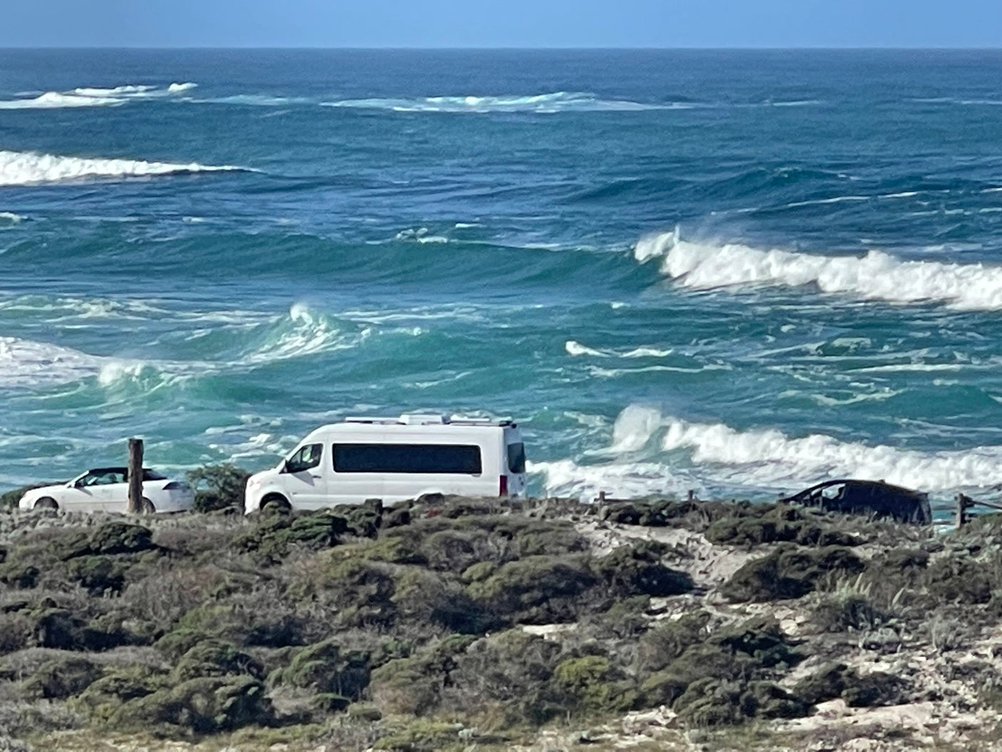Scenic coastal view: an RV rental and other vehicles parked on a bluff overlooking the ocean with breaking waves.