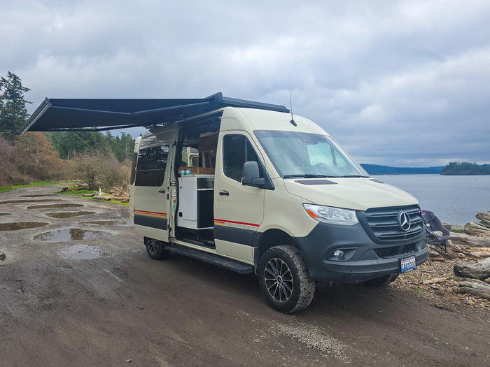 Spacious RV rental van with awning extended, parked on a muddy road by the water. Interior visible for comfortable van life.