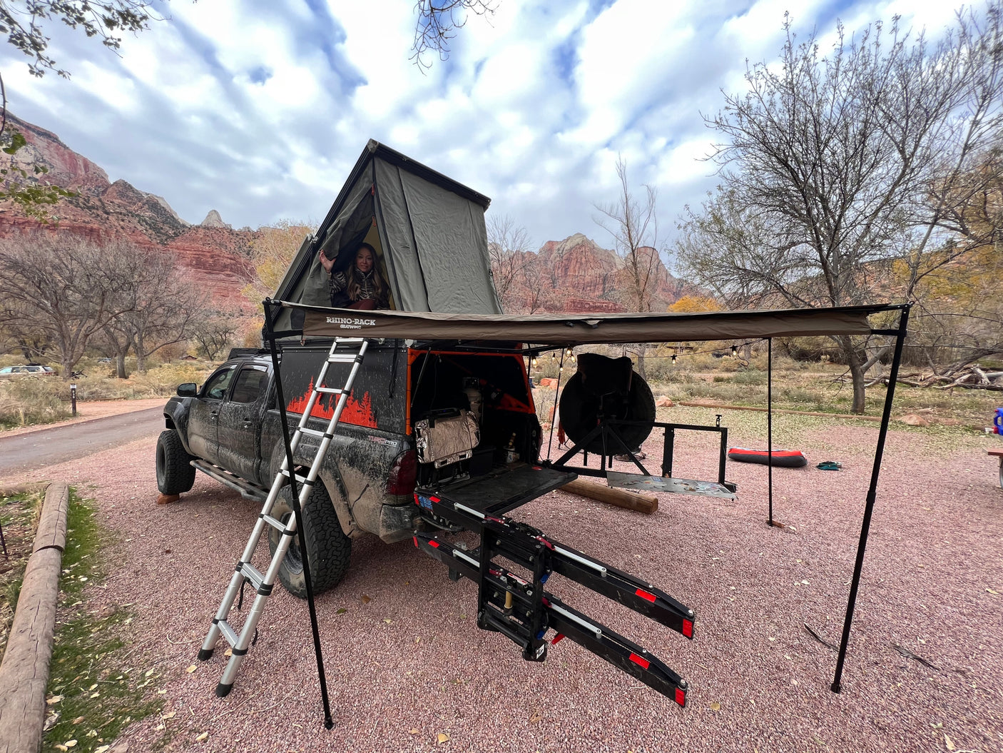 Truck with rooftop tent and awning deployed at a scenic mountain campsite. Person waving from tent. Explore the outdoors like an RV rental.